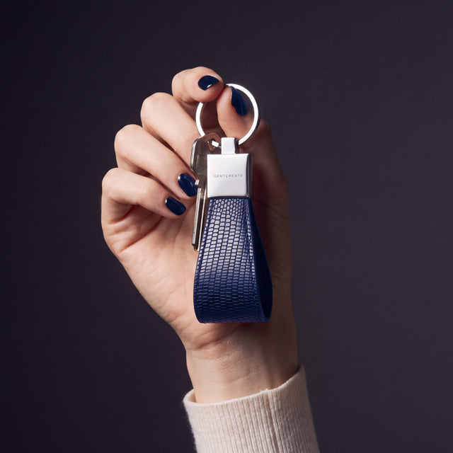 Woman's hand holding up a navy lizard-embossed leather keychain by GENTCREATE by its split ring in a dark studio