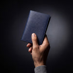 Man's hand holding a navy lizard-embossed leather passport holder by GENTCREATE in a dark studio