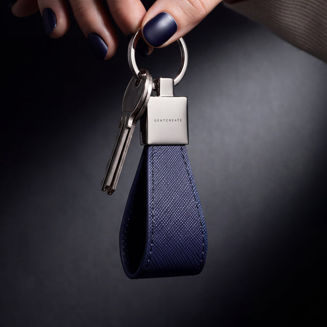 Woman's hand holding up a navy Saffiano leather keychain by GENTCREATE by its split ring in a dark studio