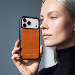 窓からの自然光の中で、GENTCREATEのリザード型押しカーフレザー iPhone 18 Pro Maxケース（オレンジ）を顔の横で持つ女性