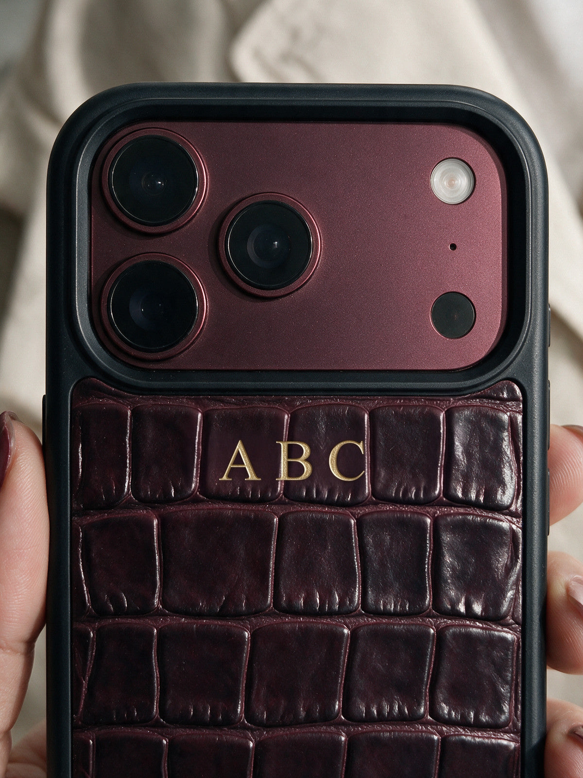Visualization: a hand holds a burgundy croco-embossed calfskin iPhone case on day 1. The initials ABC are hot-stamped by hand in 24-karat gold foil below the camera cut-out.