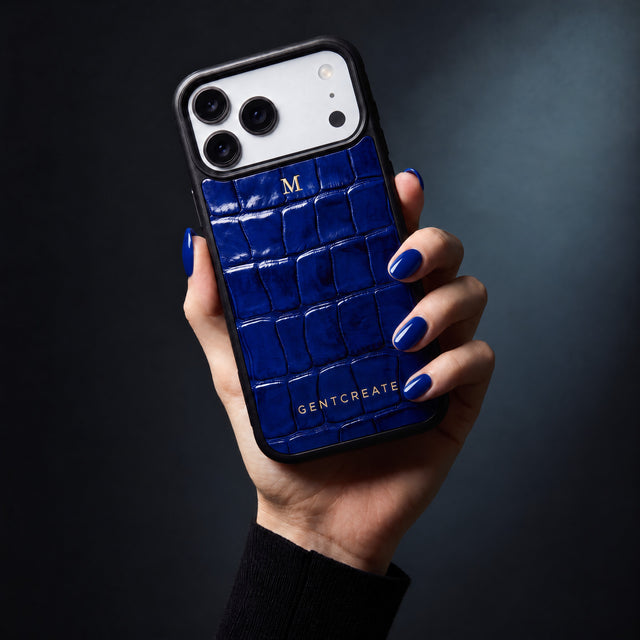 يد امرأة تمسك جرابًا أزرق لهاتف iPhone 18 Pro Max بنقش التمساح البارز من GENTCREATE يحمل الحرف الأول M بختم حراري ذهبي