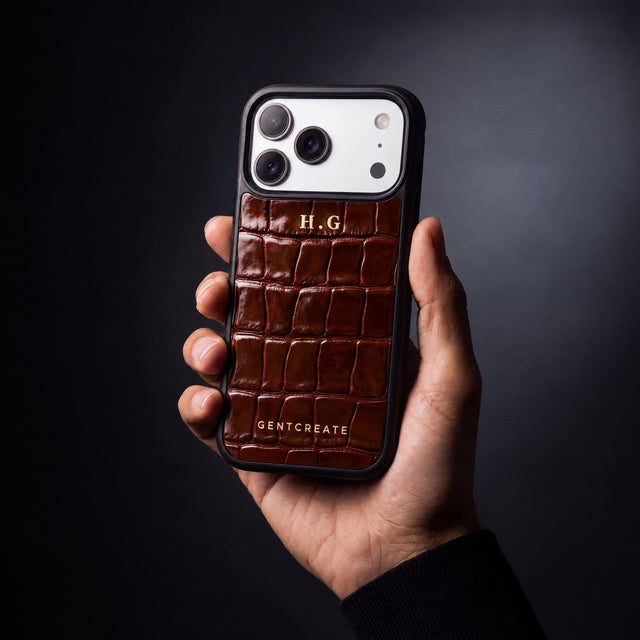 يد رجل تمسك جرابًا بنيًا لهاتف iPhone 18 Pro Max بنقش التمساح البارز من GENTCREATE يحمل الحرفين الأولين H.G بختم حراري ذهبي
