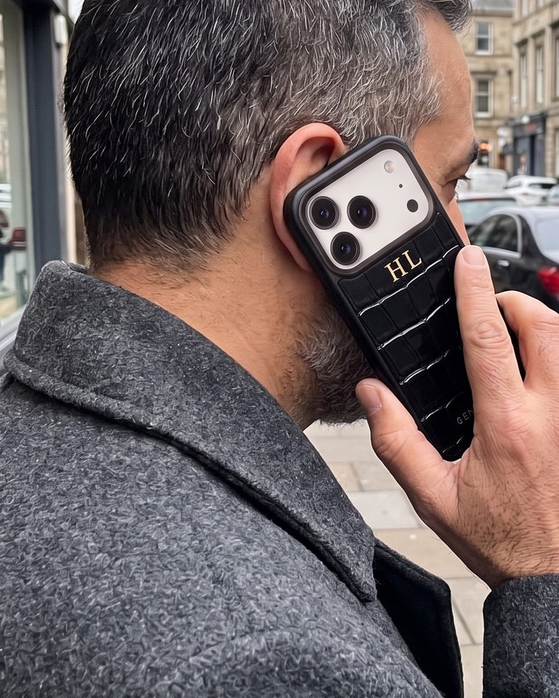 Seen from behind his shoulder on a city street, a man holds his phone to his ear in a black croco-embossed leather case with the initials HL hot-stamped in gold