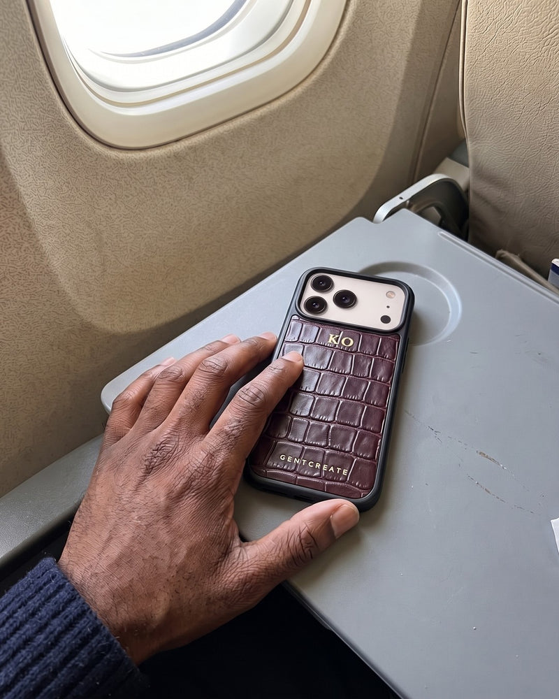 A man's hand rests on a burgundy croco-embossed leather iPhone case with the initials KO hot-stamped in gold, lying on an airplane tray table by the window