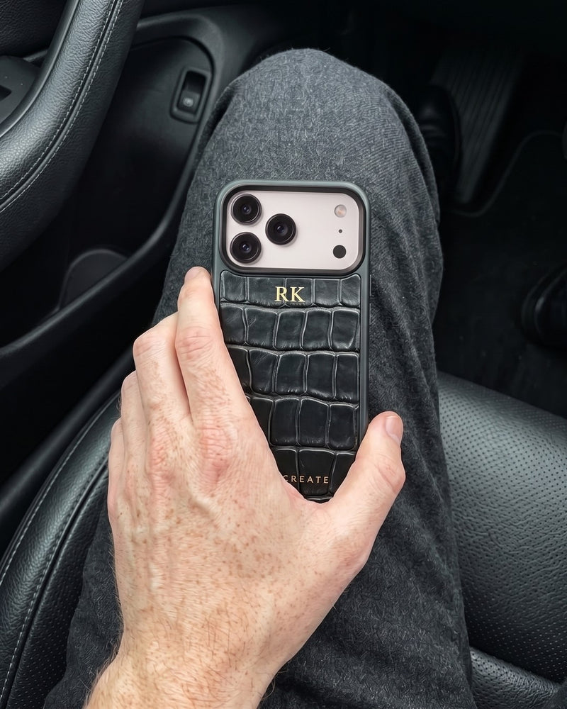 Looking down at his lap in a car, a man holds his phone in a black croco-embossed leather case with the initials RK hot-stamped in gold