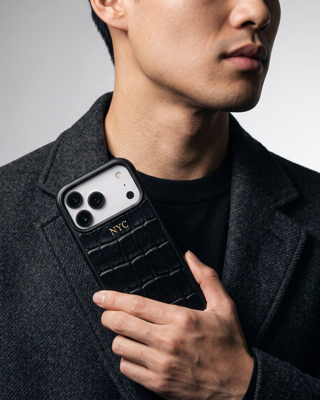 In a gray studio, a man in a charcoal wool coat holds his phone against his chest in a black croco-embossed leather case hot-stamped NYC in gold