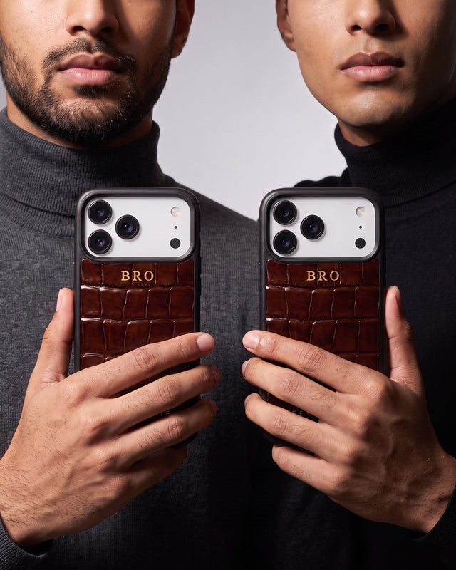 In a gray studio, two young men in dark turtlenecks stand side by side holding up their phones in matching brown croco-embossed leather cases, each hot-stamped BRO in gold