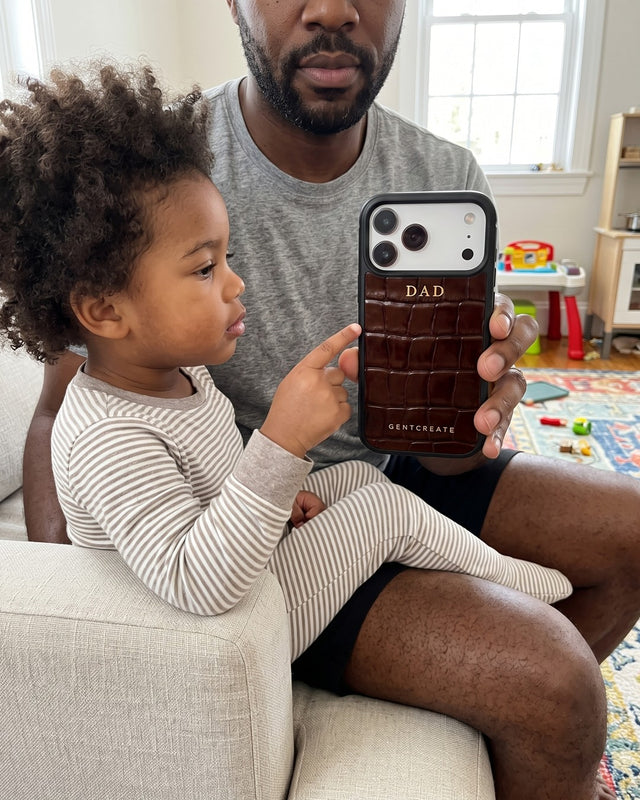 A father on a sofa holds up his phone in a brown croco-embossed leather case hot-stamped DAD in gold while his young daughter points at the letters