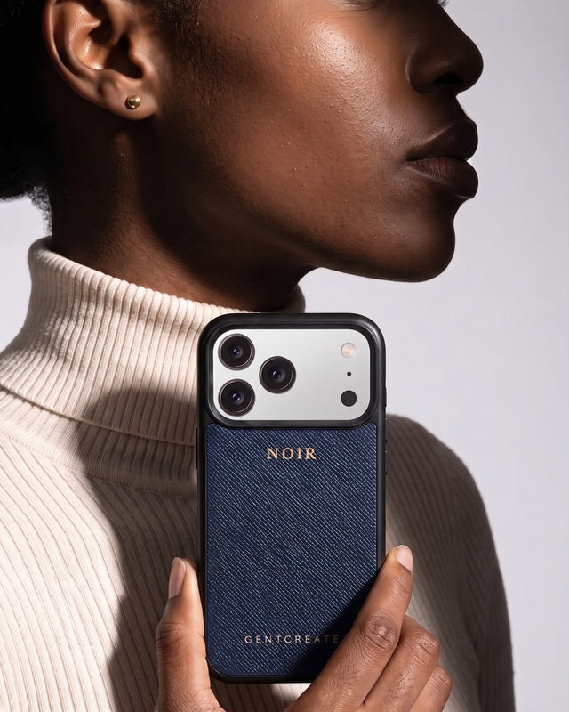 In a gray studio, a woman in a cream ribbed turtleneck, seen in profile, holds up her phone in a navy saffiano leather case hot-stamped NOIR in gold