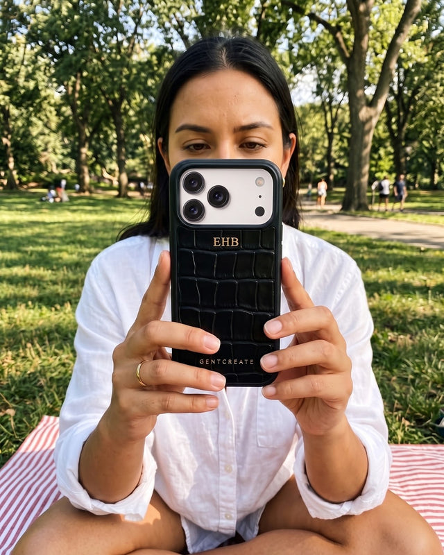 A woman sitting on a picnic blanket in a park holds up her phone in a black croco-embossed leather case hot-stamped in gold with the initials EHB