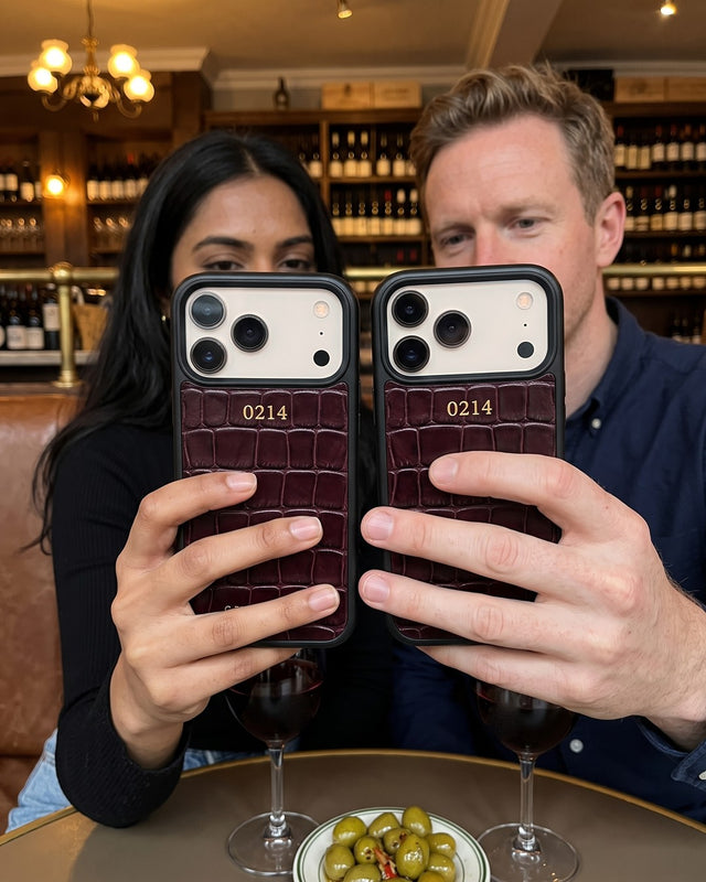 A couple at a wine bar hold up their phones side by side in matching burgundy croco-embossed leather cases, each hot-stamped in gold with 0214