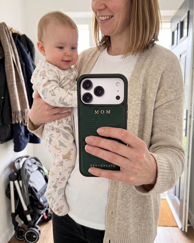 A mother holding her baby on her hip in the front hallway shows her phone in a green lizard-embossed leather case hot-stamped MOM in gold