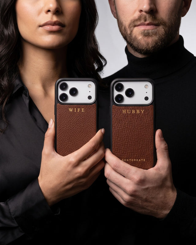 In a gray studio, a couple in black stand shoulder to shoulder holding up their phones in matching brown pebble-grain leather cases, one hot-stamped WIFE and one HUBBY in gold
