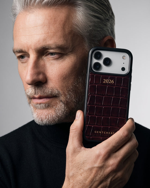 In a gray studio, a silver-haired man in a black turtleneck holds his phone to his ear, its burgundy croco-embossed leather case hot-stamped 2026 in gold
