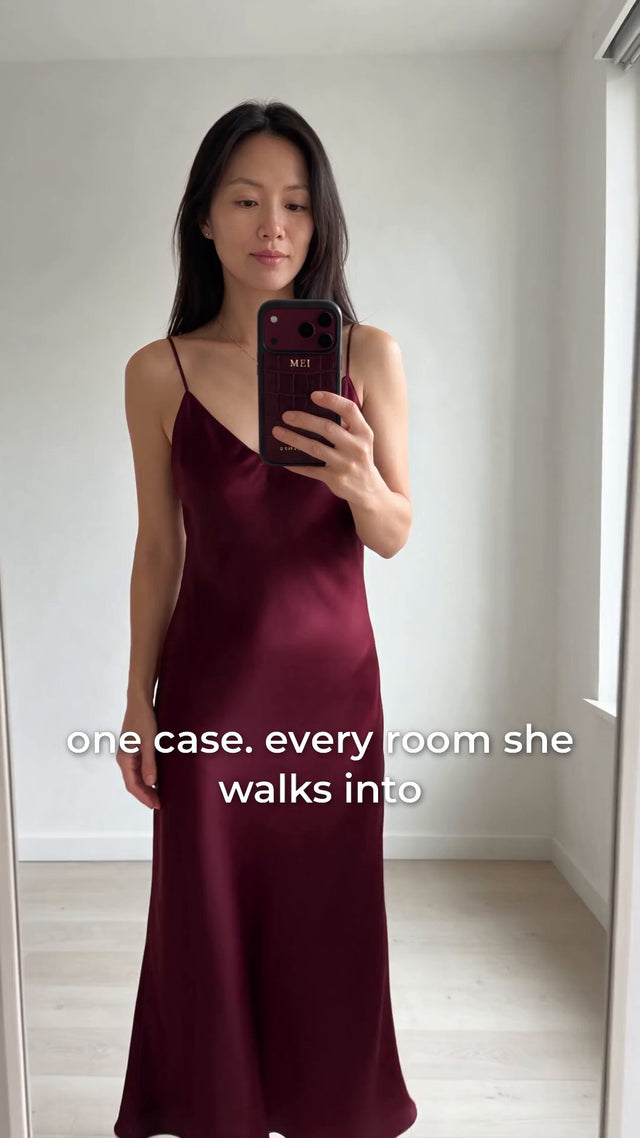 A woman in a burgundy satin dress takes a mirror selfie with a burgundy croco-embossed leather iPhone case hot-stamped in gold