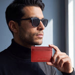 Uomo con occhiali da sole che tiene un portacarte GENTCREATE in pelle rossa goffrata a onde sotto il mento, alla luce di una finestra