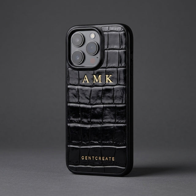 Black croco-embossed glossy leather iPhone case with the initials AMK hot-stamped in gold, standing at an angle on charcoal