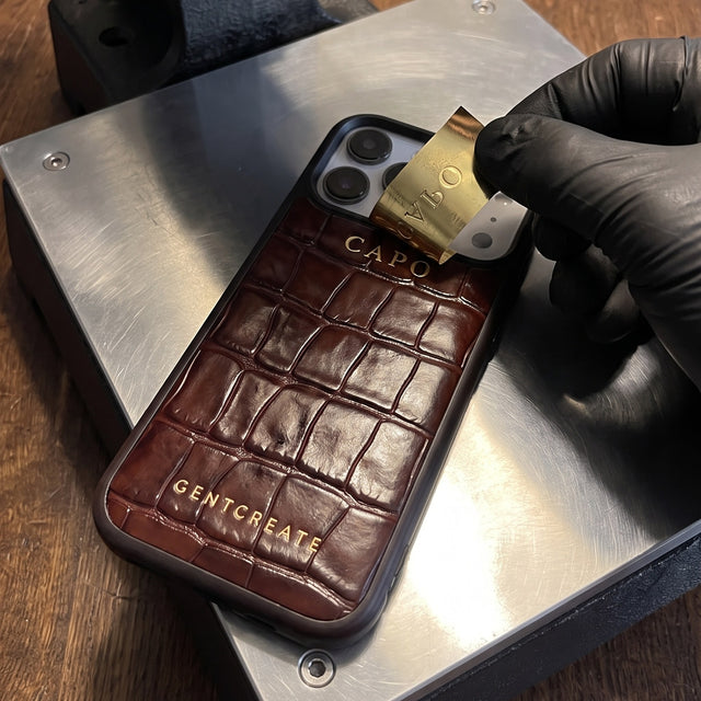 Gloved hand peeling gold foil off the freshly hot-stamped word CAPO on a brown croco-embossed leather iPhone case