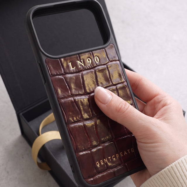 Burgundy croco-embossed glossy leather iPhone case with the initials LN90 hot-stamped in gold, held in front of an open black GENTCREATE box