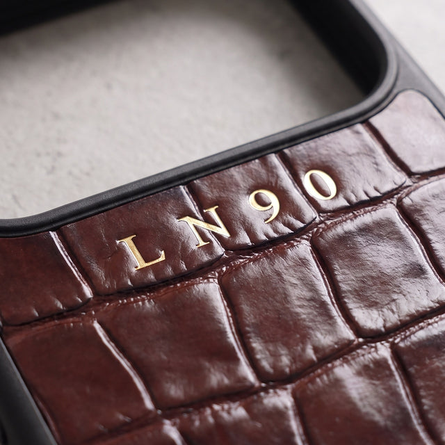 Close-up of the initials LN90 hot-stamped in gold foil on burgundy-brown croco-embossed glossy leather, just below the camera opening