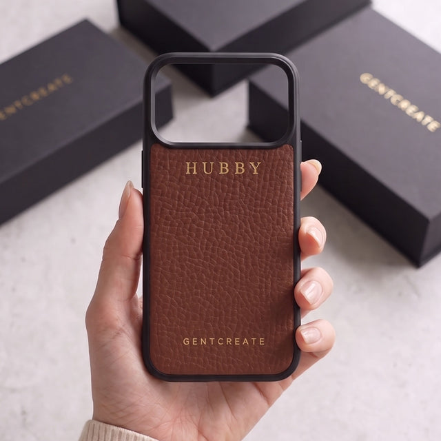 Brown pebble-grain leather iPhone case with HUBBY hot-stamped in gold, held in hand in front of black GENTCREATE boxes